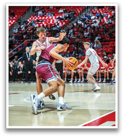 A basketball game is in progress with players on the court. One player is attempting to block a shot, while another player is trying to get the ball. The players are wearing uniforms and are in the middle of the action. AI generated content