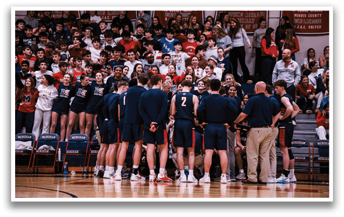 A group of young men are standing on a basketball court, some wearing uniforms. They are gathered together, possibly discussing their game strategy. The crowd of spectators is watching the game intently, filling the stands and surrounding the court. AI generated content