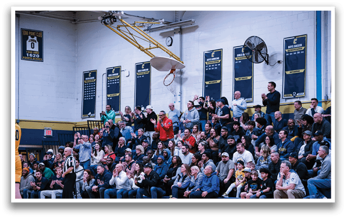 A basketball game is taking place in a gymnasium, with a crowd of people watching the game. The spectators are seated on benches, and some are standing. The atmosphere is lively, with people cheering and clapping for the players. The gymnasium has a clock on the wall, and a fan is present to help circulate air. The crowd is diverse, with people of different ages and genders, all gathered to enjoy the game. AI generated content
