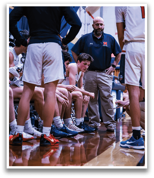 A group of men are sitting on the floor, with one man standing over them. The standing man is wearing a blue shirt and appears to be a coach. The men sitting on the floor are wearing uniforms, suggesting they are part of a sports team. AI generated content