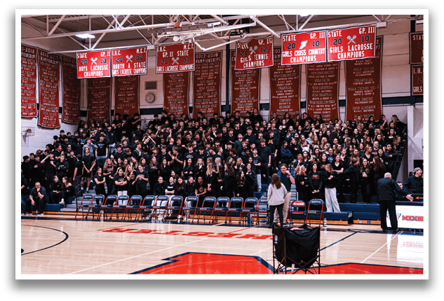 A large crowd of people is gathered in a gymnasium, sitting on chairs and standing around. The audience is watching a game or an event, and there are several clocks visible in the room. The people are dressed in black, and some are wearing ties. The atmosphere is lively and energetic, with everyone engaged in the ongoing activity. AI generated content