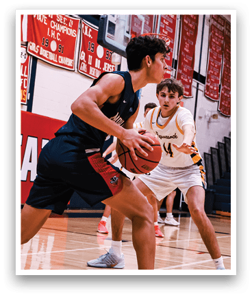 A basketball game is taking place in a gymnasium with several players on the court. One player is holding a basketball while another player is trying to block him. The players are wearing uniforms and are actively engaged in the game. AI generated content