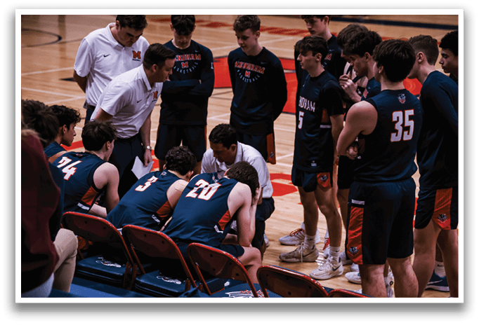 A group of young men are gathered on a basketball court, with some sitting on chairs and others standing. They are engaged in a conversation, possibly discussing their game strategy. The scene takes place in a gym, with a bench visible in the background. AI generated content