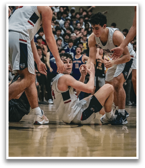 Three basketball players are on the floor, with one player holding another player's leg. The players are wearing uniforms and are surrounded by a crowd of people. AI generated content