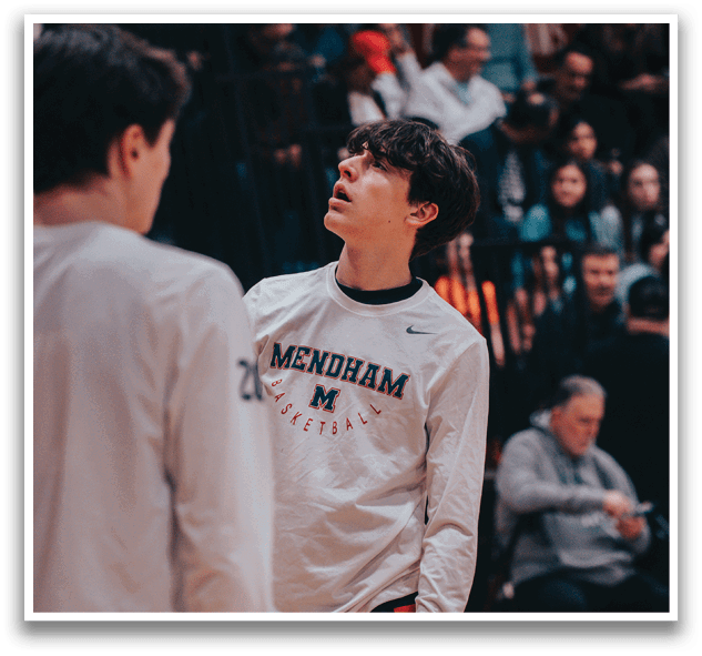 Two men wearing white shirts are standing on a basketball court. One of the men is looking up, possibly at the crowd or the scoreboard. There are several people in the background, some of whom are also wearing white shirts. A chair is visible in the background, and a handbag can be seen on the ground. AI generated content