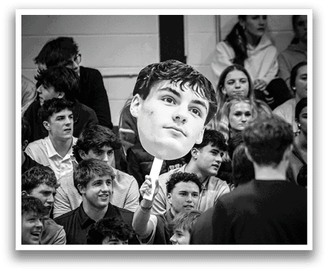 A group of people are sitting in the bleachers, watching a man holding a giant cutout of his face. AI generated content