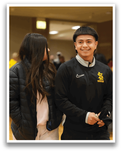 A man and woman are standing in a room, both wearing black jackets. The man is smiling and holding a remote control. There are several other people in the room, some of whom are also holding remotes. A chair is visible in the background. Contenido generado con IA