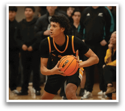 Two men are playing basketball on a court, with one of them holding a basketball. There are several other people watching the game, some of them sitting on chairs. Contenido generado con IA