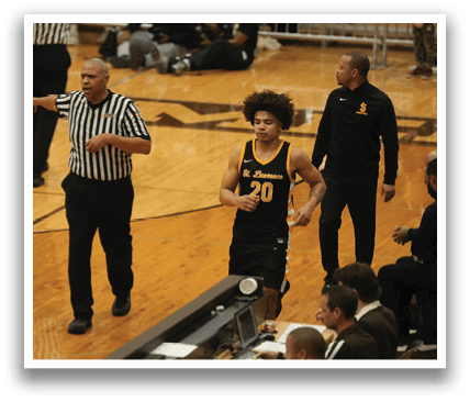 A basketball game is taking place with a referee and a player on the court. The referee is wearing a striped shirt and is holding his hand up. The player is walking by the referee. There are several other people in the scene, some of whom are sitting on chairs. Contenido generado con IA