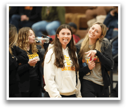 A basketball game is taking place with a referee and a player on the court. The referee is wearing a striped shirt and is holding his hand up. The player is walking by the referee. There are several other people in the scene, some of whom are sitting on chairs. Contenido generado con IA