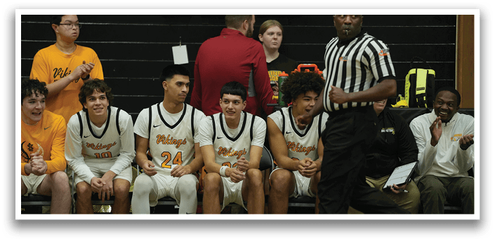 A group of basketball players are sitting on a bench, watching the game. A referee is standing nearby, overseeing the match. AI generated content