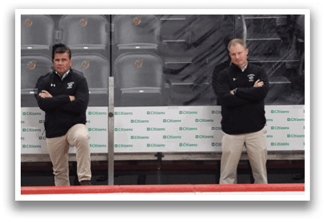 Two men are standing on the ice, wearing black and white uniforms. They are both leaning on the boards, possibly taking a break or waiting for their turn to play. AI generated content