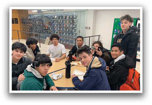 A group of teenagers are sitting around a table, eating and drinking. They have various items on the table, including a bottle, a cup, a sandwich, and a donut. Some of the teenagers are wearing backpacks, and a few of them are holding cell phones. The scene appears to be a casual gathering of friends. AI generated content