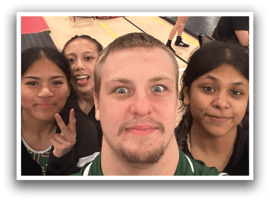 A group of people, including a man and three women, are standing in a room. The man is making a funny face and appears to be taking a selfie. The women are standing around him, and there is a chair in the background. AI generated content