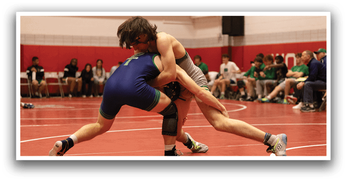 Two wrestlers are grappling on a red mat, with one of them wearing a blue and green outfit. The wrestlers are surrounded by a crowd of spectators, including people sitting on chairs and others standing. The wrestlers are in the center of the action, with their bodies entwined as they compete. AI generated content
