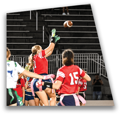 A person in football gear and helmet is crouched on a field with other players lined up in the background during a game. Description generated by AI