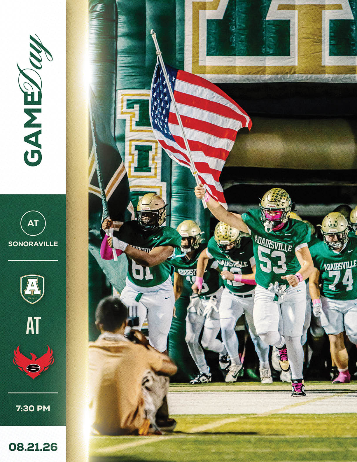 A group of football players in green and white uniforms run onto the field with one player holding an American flag while a person in tan clothing sits on the ground nearby. Description generated by AI