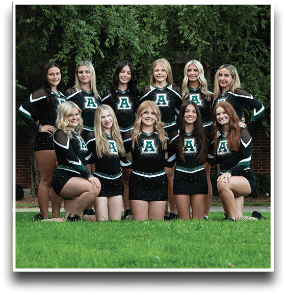 A group of eleven people wearing matching black and green cheerleading uniforms with the letter A on the front are posing outdoors on grass in front of a brick wall and trees. Description generated by AI