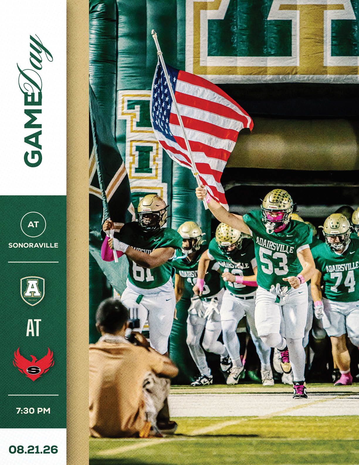 People in football uniforms run onto the field from a tunnel, with one person holding an American flag, while a photographer kneels on the sidelines capturing the moment. Description generated by AI