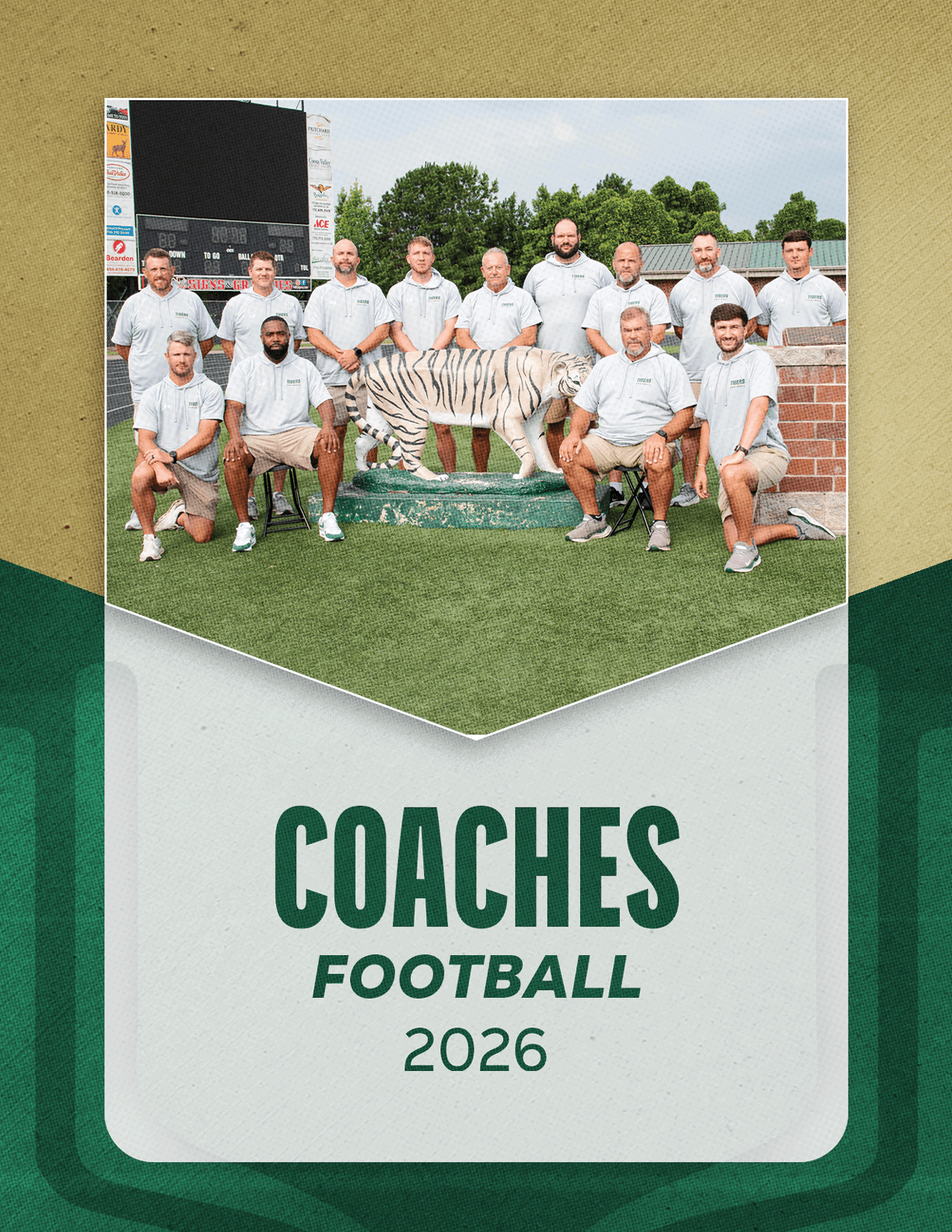A group of adults wearing matching light-colored polo shirts pose outdoors on a sports field around a white tiger statue with the text coaches football 2026 below. Description generated by AI