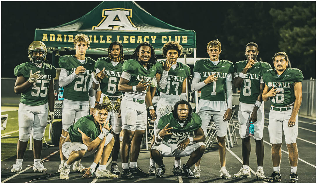 A group of football players in green and white uniforms pose together on a field in front of a tent labeled Adairsville Legacy Club at night. Description generated by AI