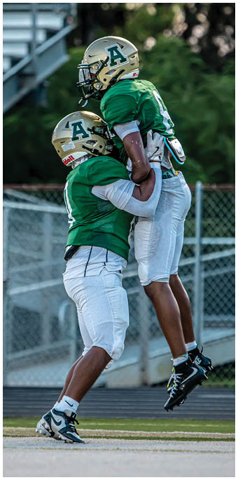 Two people wearing green sports jerseys and gold helmets with a large letter A are positioned side by side near bleachers. Description generated by AI