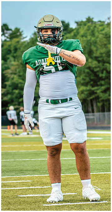 A person wearing a green football jersey and white pants stands on a football field with trees in the background. Description generated by AI
