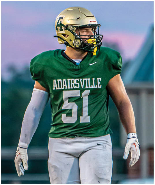 A person wearing a green football jersey with the number 51 and a gold helmet is standing on a field during a game at dusk. Description generated by AI