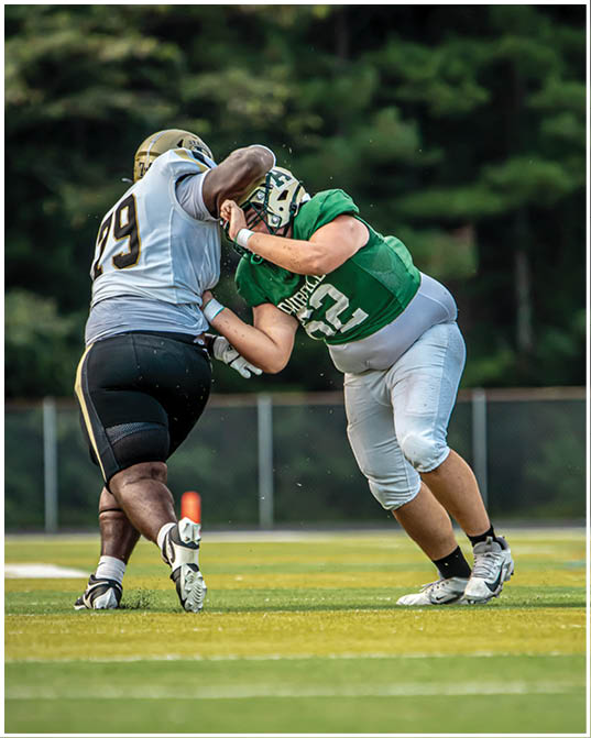 Two people in football uniforms are engaged in a tackle on a grassy field with a blurred fence in the background. Description generated by AI