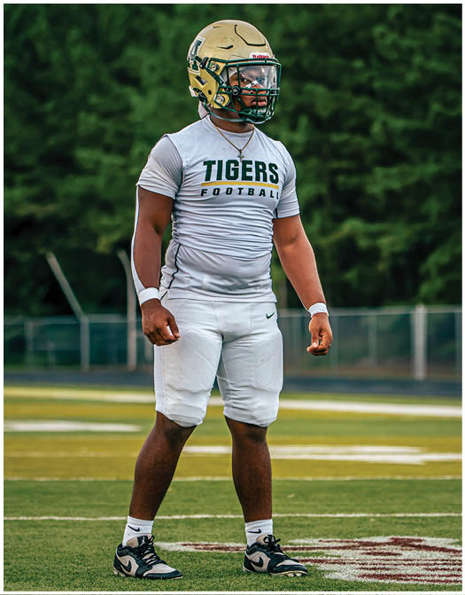 A person wearing a green sports jersey with the number 10 is adjusting a gold football helmet against a blue sky background. Description generated by AI