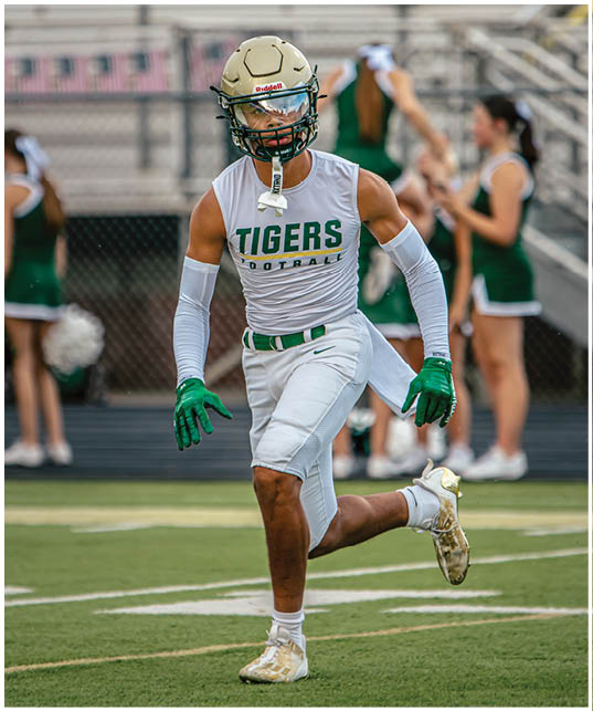 A person wearing a football helmet, green gloves, and a white sleeveless shirt with “Tigers Football" printed on it is running on a sports field with cheerleaders in green uniforms blurred in the background. Description generated by AI