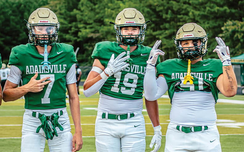 Three people wearing green football uniforms and helmets stand side by side outdoors with trees in the background, each holding up two fingers. Description generated by AI