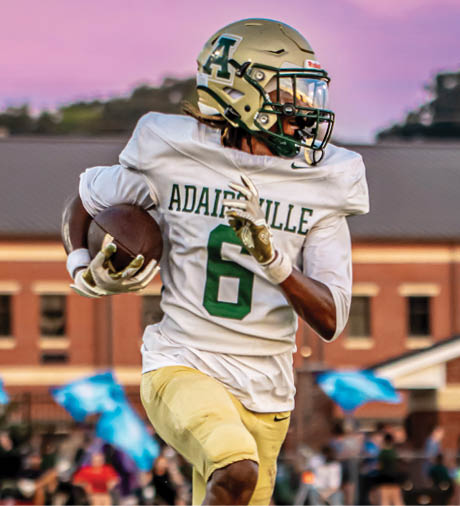 A person wearing a football uniform with the number six is running on a grassy field while holding a football, with a building and spectators in the background. Description generated by AI