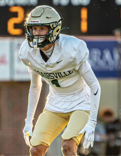 A person in a football uniform and helmet is standing in a ready position on a sunlit field with a scoreboard in the background. Description generated by AI