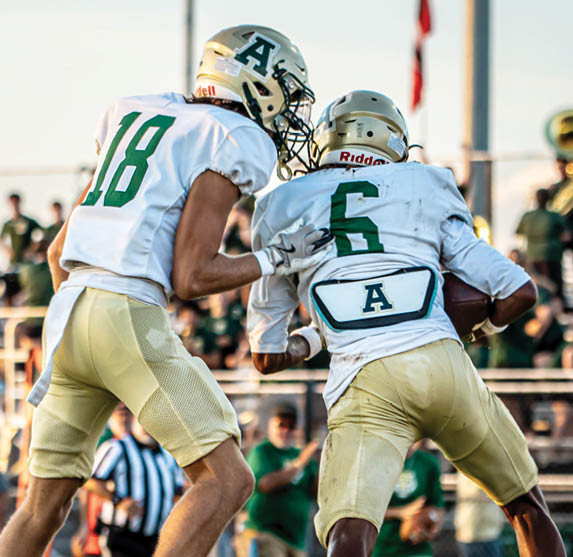 Two people in football uniforms with helmets are engaged in a play on a field, with one holding a football and the other appearing to assist or block. Description generated by AI