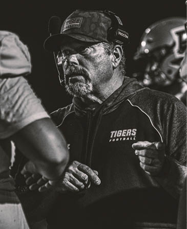 A person wearing a cap with a headset and a hoodie with the word “TIGERS" on it is looking intently to the side in a black and white photo. Description generated by AI