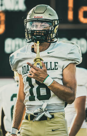 A person wearing a football uniform with the number 18 and a helmet is standing on a field with a scoreboard in the background. Description generated by AI