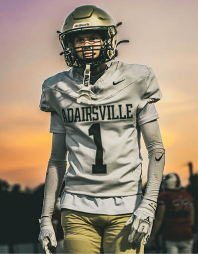 A person wearing a football helmet and a white jersey with the word “ADAIRSVILLE" printed on it is smiling. Description generated by AI