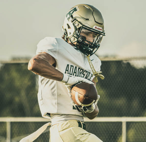 A person wearing a football uniform and helmet is holding a football while running on a field with a chain-link fence and trees in the background. Description generated by AI
