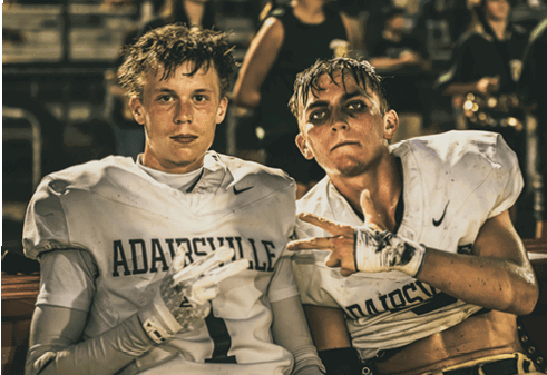 Two people wearing football uniforms with the name “Adairsville" are sitting close together, one making a peace sign with their hand. Description generated by AI