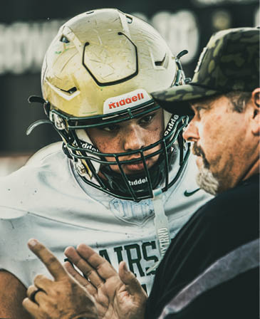 A person wearing a football helmet and jersey is closely listening to another person who is speaking and gesturing with their hand. Description generated by AI