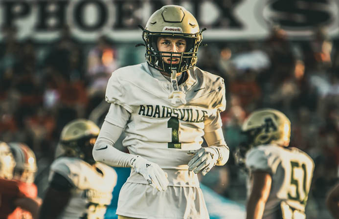 A person wearing a football uniform with the number one stands on a field with two other players in the background during a game. Description generated by AI