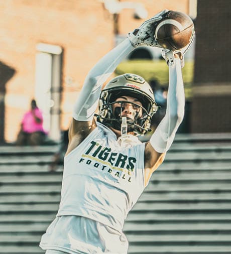 A person wearing a football uniform with the number six is running on a grassy field while holding a football, with a building and spectators in the background. Description generated by AI