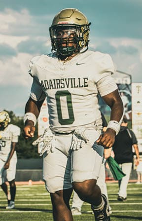 A person wearing a football uniform with the number 18 and a helmet is standing on a field with a scoreboard in the background. Description generated by AI