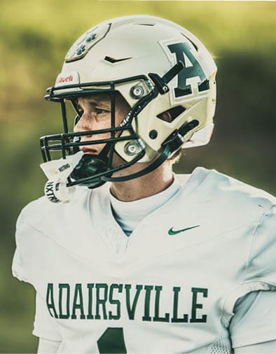 A person wearing a football helmet and a white jersey with the word “ADAIRSVILLE" printed on it is smiling. Description generated by AI