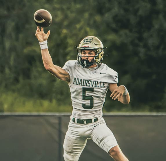 A person wearing a football uniform and helmet is holding a football while running on a field with a chain-link fence and trees in the background. Description generated by AI