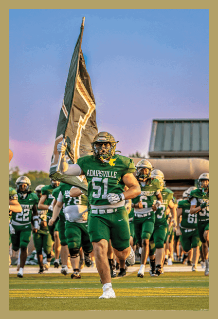 A person in a green football uniform with the number 51 is running on a field holding a large flag while followed by a group of people in similar uniforms. Description generated by AI