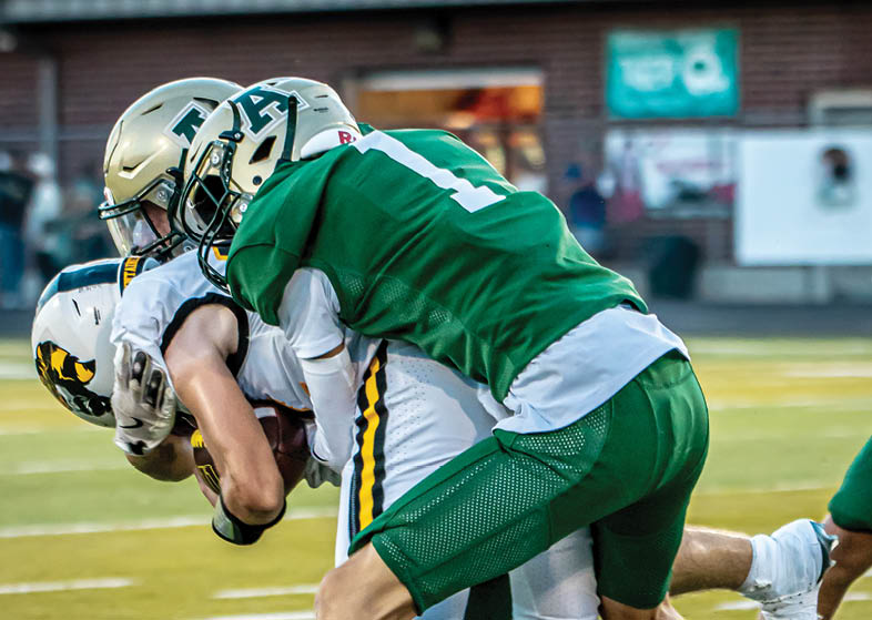 Two people in green football uniforms tackle a person in a white and yellow football uniform on a field during a game. Description generated by AI