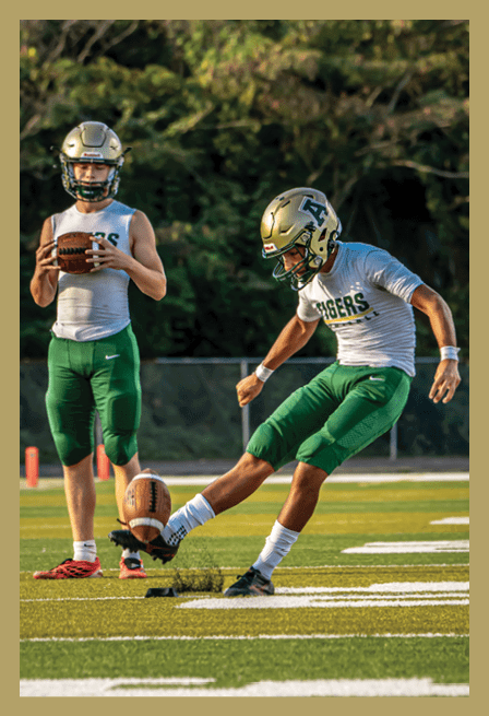 A person in a green football uniform with the number 51 is running on a field holding a large flag while followed by a group of people in similar uniforms. Description generated by AI