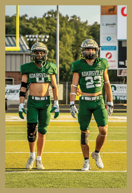 Two adults wearing green football uniforms and helmets walk side by side on a football field during daylight. Description generated by AI